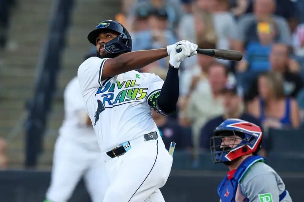 A baseball player in a blue and white jersey swings a bat during a game, with an audience visible in the background.