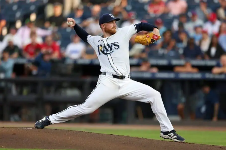 A pitcher in a white "Rays" jersey prepares to throw a baseball on the mound, with a crowd of fans visible in the background.