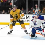 A hockey game scene with a player in a gold jersey preparing to take a shot, while the goalie in blue and orange braces for action.