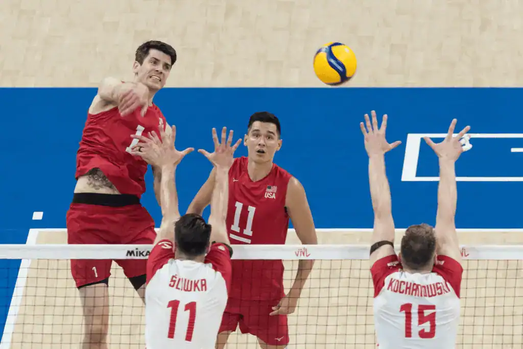 Una escena de un partido de voleibol que muestra a jugadores de EE. UU. y Polonia saltando para bloquear una pelota que vuela alto sobre la red.