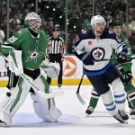A tense hockey game scene featuring a goalie in green gear, a player in white with blue accents, and action taking place near the net.