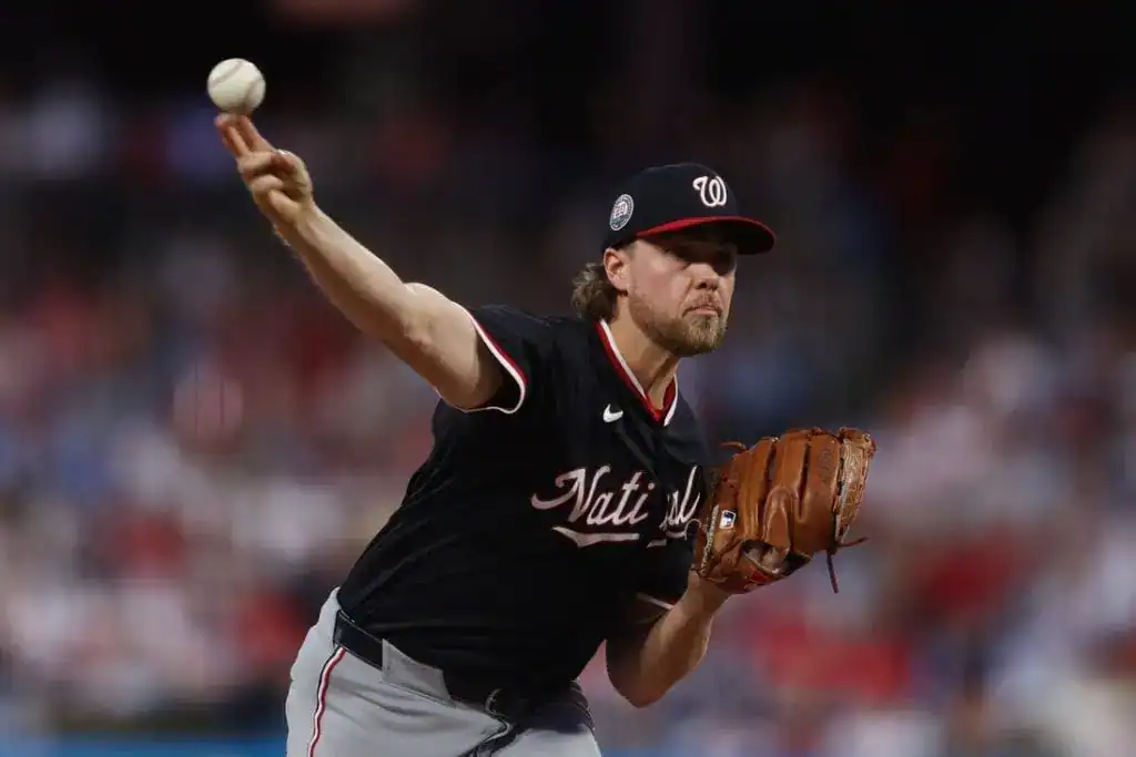 A pitcher in a black Washington Nationals jersey throws a baseball, focused on the play amid a blurred stadium background.