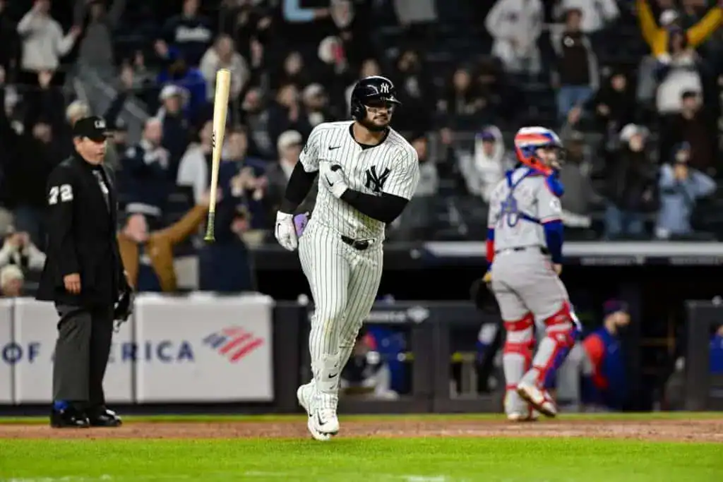 A New York Yankees player runs enthusiastically after hitting the ball, with fans cheering in the stands behind him.