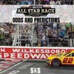 A NASCAR car crosses the finish line at North Wilkesboro Speedway, surrounded by cheering fans and a banner for the 2025 All-Star Race.