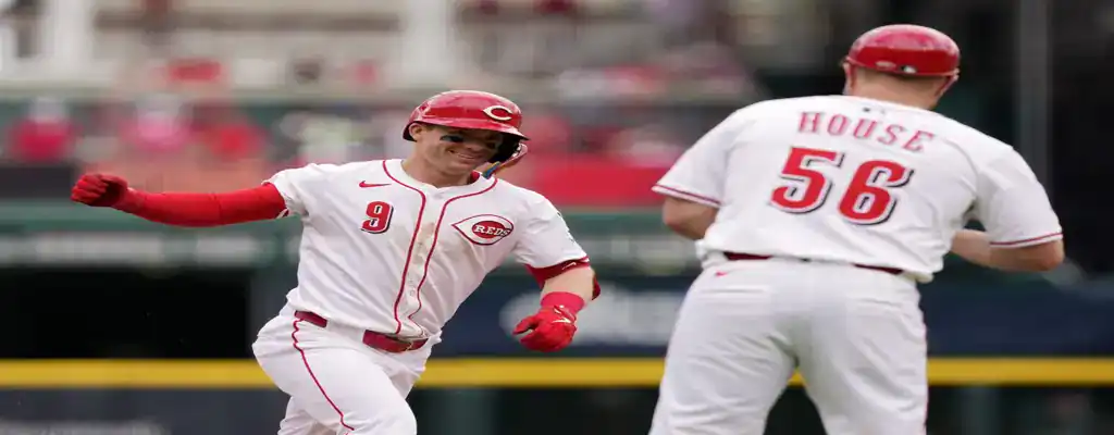 Cincinnati Reds player smiles and celebrates while rounding the bases during a baseball game, wearing a white uniform with red details.