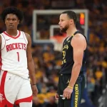 Two basketball players face each other on the court, one in a red Houston Rockets jersey and the other in a black Golden State Warriors jersey.