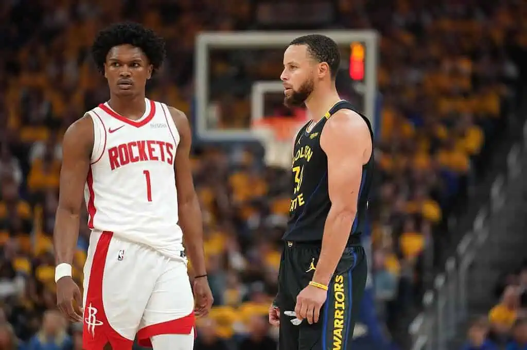 Two basketball players face each other on the court, one in a red Houston Rockets jersey and the other in a black Golden State Warriors jersey.