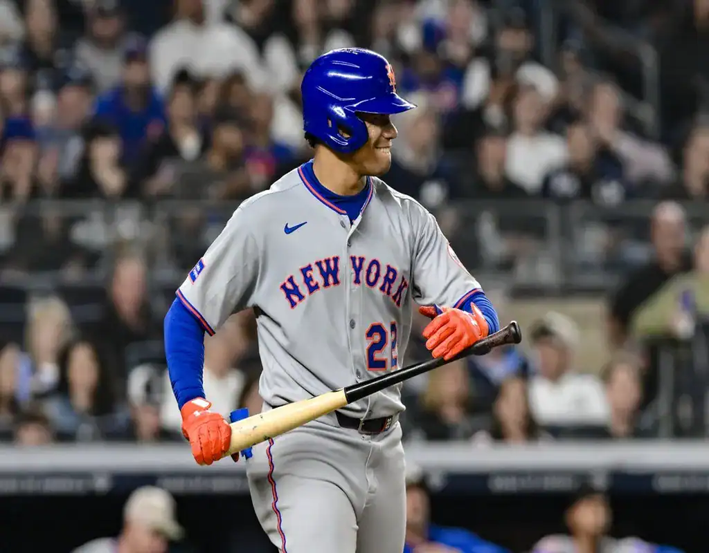 New York Mets batter smiling and holding his bat in the on-deck circle with a packed stadium behind him