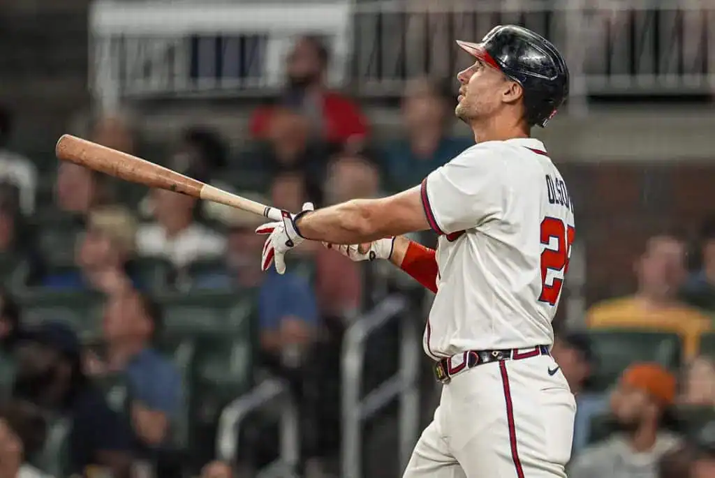 A baseball player in a white jersey with red accents swings a bat in a stadium filled with spectators, capturing the action of a game.