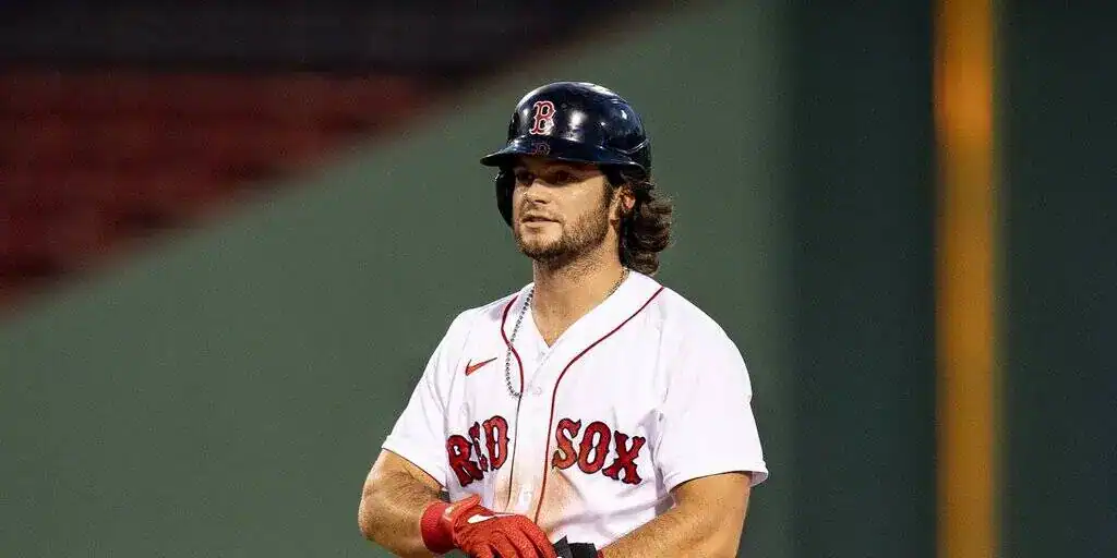 A Boston Red Sox player stands on the field, wearing a white jersey and a helmet, ready for play at a baseball stadium.