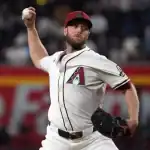 A baseball pitcher in a white Arizona Diamondbacks jersey prepares to throw a ball, with a vibrant crowd and stadium setting behind him.