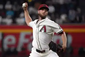A baseball pitcher in a white Arizona Diamondbacks jersey prepares to throw a ball, with a vibrant crowd and stadium setting behind him.