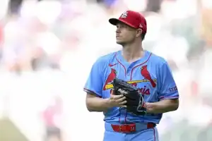 A baseball player in a light blue jersey with red accents stands on the field, holding a glove and preparing for play.