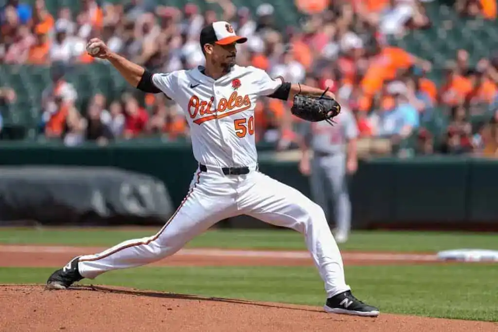 A baseball pitcher in an orange and white Orioles uniform winds up to throw a pitch on a sunny day at the stadium, with fans in the background.