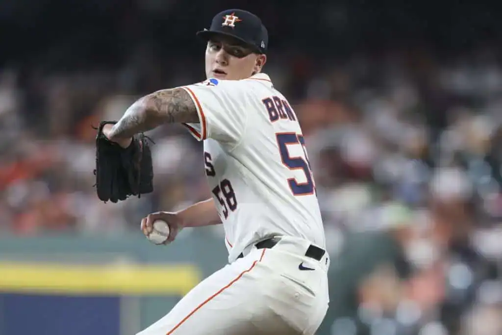 A baseball player in an orange and white jersey is pitching on the field, with a crowd blurred in the background.