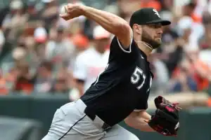 A baseball pitcher in a black jersey throws a pitch on the mound, surrounded by an enthusiastic crowd in the background.