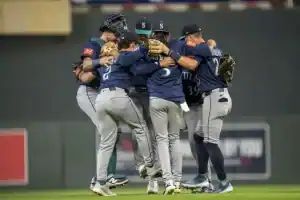 A group of baseball players in Mariners jerseys joyfully celebrates in a tight huddle on the field after a significant play.
