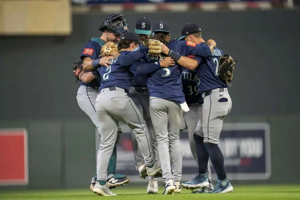 A group of baseball players in Mariners jerseys joyfully celebrates in a tight huddle on the field after a significant play.