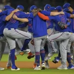 A group of baseball players in blue jerseys huddle closely, celebrating in a lively, interconnected pose on the field.