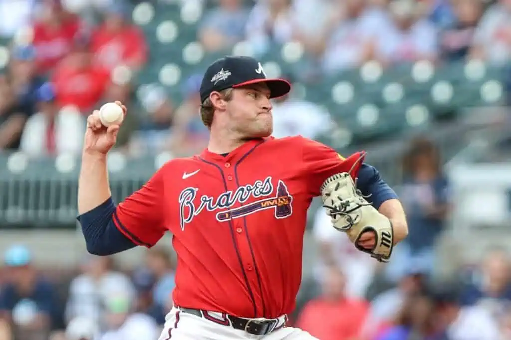 A baseball player in a red Atlanta Braves jersey prepares to pitch, holding a ball, with a stadium full of fans in the background.