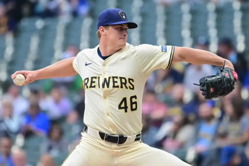 A baseball player in a Brewers jersey winds up to pitch on the mound, surrounded by a cheering crowd in a stadium.