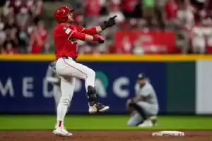 A baseball player in a red uniform celebrates enthusiastically after hitting a play, with a cheering crowd in the background.