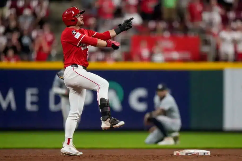 A baseball player in a red uniform celebrates enthusiastically after hitting a play, with a cheering crowd in the background.