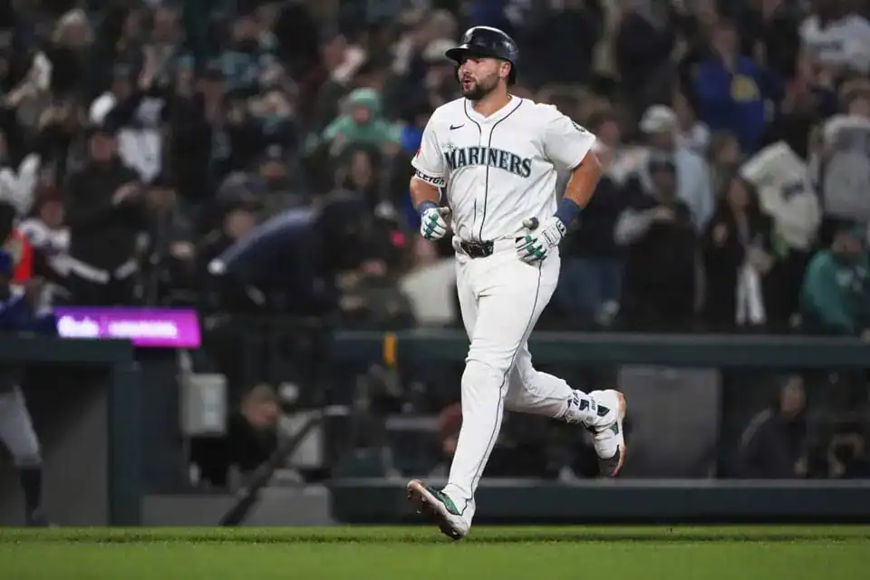 A baseball player in a Mariners uniform jogs across the field, surrounded by a cheering crowd at a nighttime game.