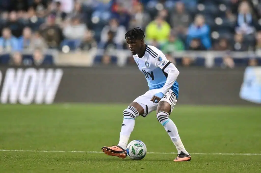 A soccer player in a light blue and white jersey is in an action pose, preparing to kick the ball on a stadium field.