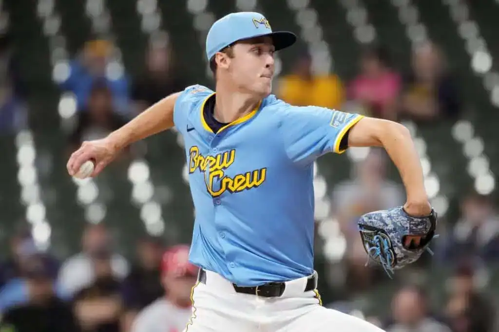 A baseball player in a light blue jersey with "Brew Crew" lettering throws a pitch on the field, surrounded by spectators.