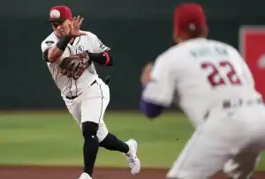 A player in a white baseball uniform makes a catch while another teammate watches closely. The field is a vibrant green background.
