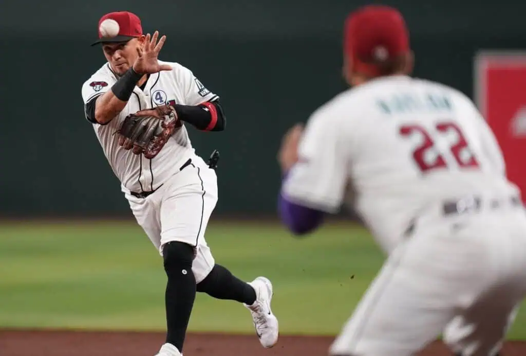 A player in a white baseball uniform makes a catch while another teammate watches closely. The field is a vibrant green background.