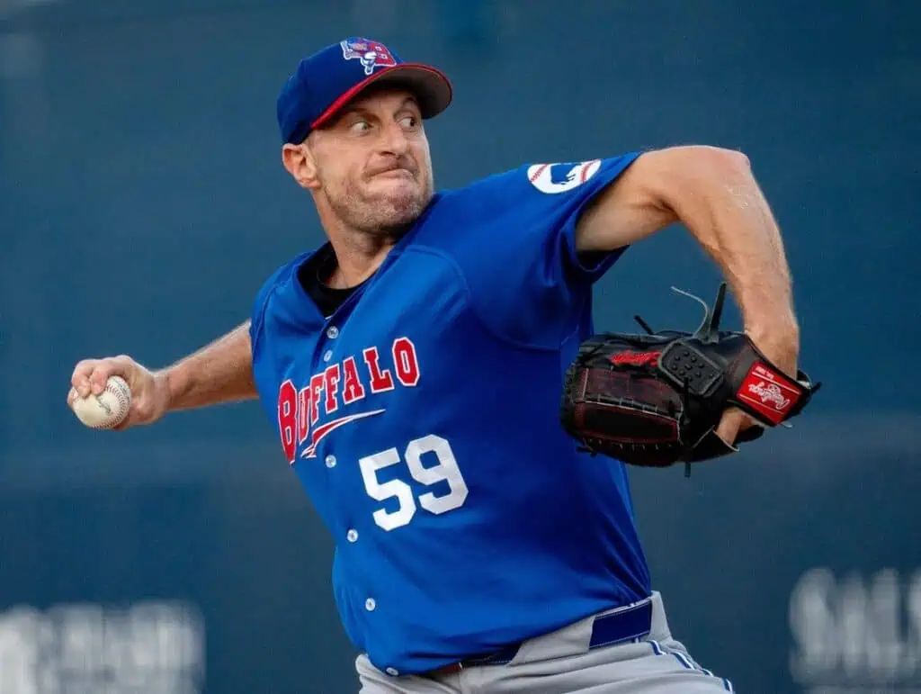 A baseball pitcher in a blue team jersey with 'Buffalo' emblazoned throws a pitch, gripping a baseball in his right hand.