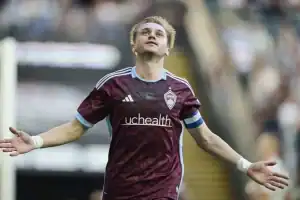 A soccer player in a maroon jersey with light blue accents celebrates a goal, arms outstretched, in front of a cheering crowd.