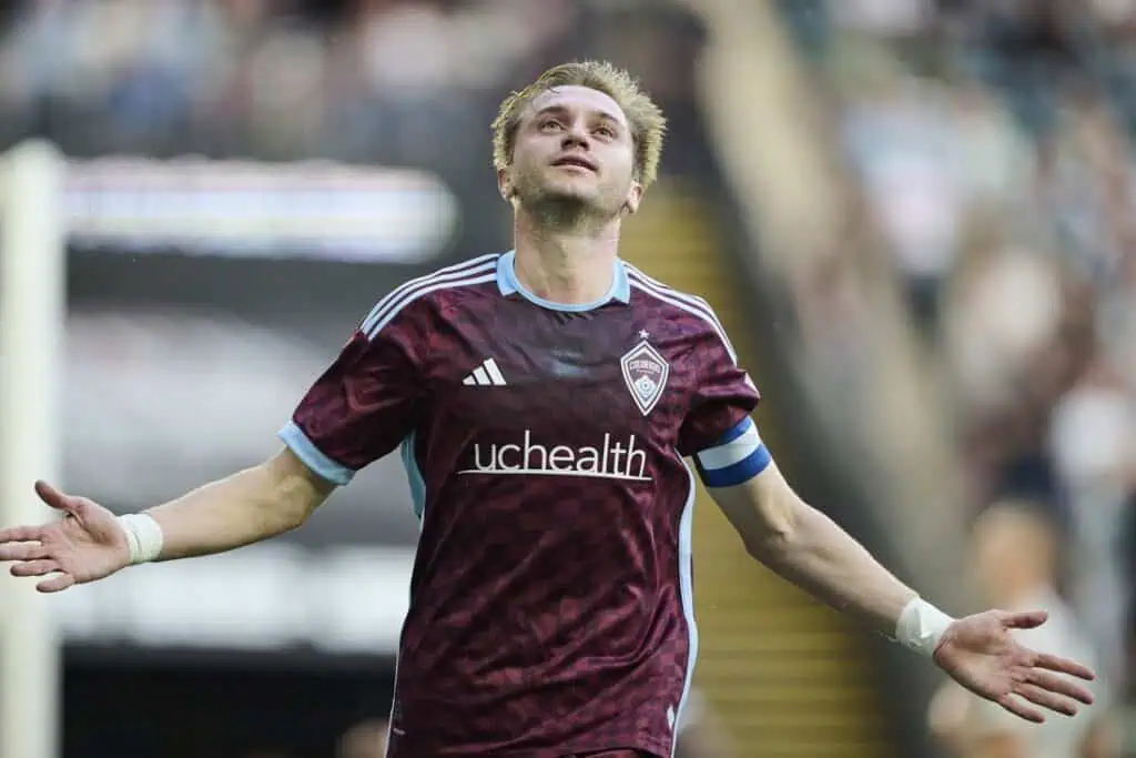 A soccer player in a maroon jersey with light blue accents celebrates a goal, arms outstretched, in front of a cheering crowd.