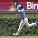 A baseball player in a blue jersey sprints towards home plate, arms raised in celebration against a green ivy backdrop.