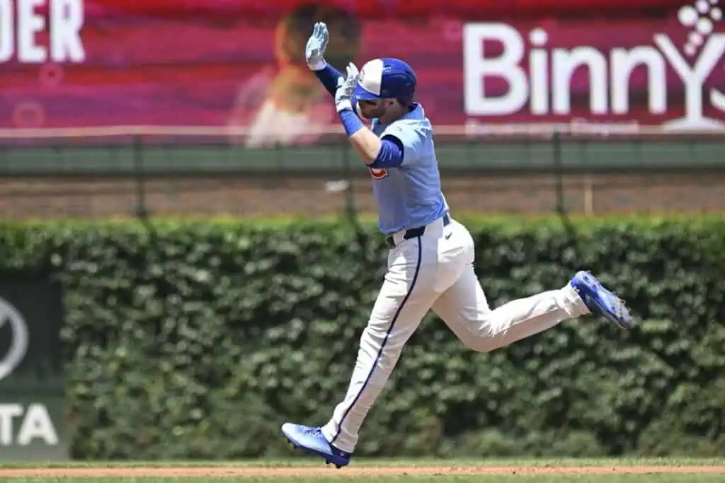 A baseball player in a blue jersey sprints towards home plate, arms raised in celebration against a green ivy backdrop.