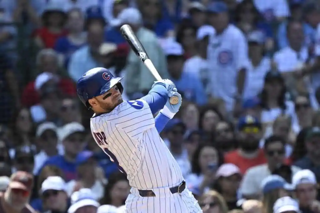 A baseball player in a Cubs uniform swings a bat at home plate during a game, surrounded by a cheering crowd in the stands.