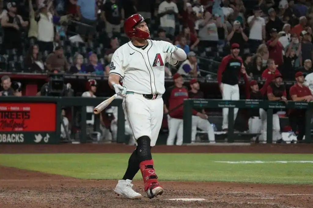 A baseball player in a white uniform stands at home plate, bat raised, while fans celebrate in the background during a night game.