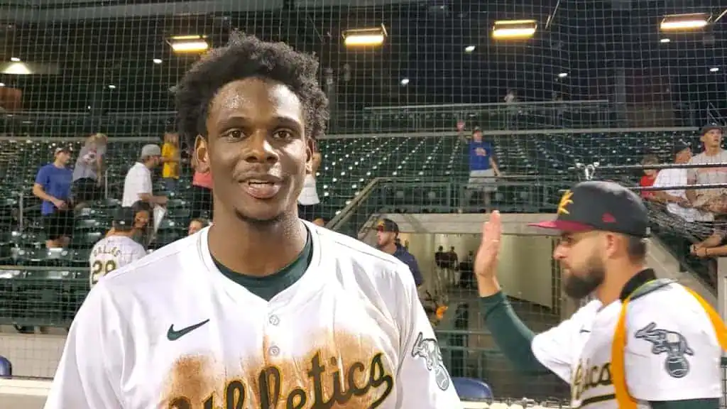 Los Angeles Angels vs Oakland Athletics Picks and Predictions June 10th 2025 2 A baseball player in a white jersey with "Athletics" printed, stands smiling in front of an empty stadium with fans in the background.