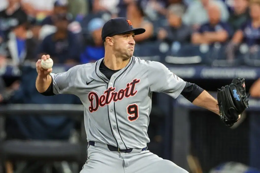 A Detroit Tigers pitcher in a gray uniform winds up to throw a baseball during a game, with fans visible in the background.
