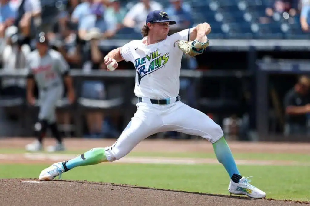 A pitcher in white and teal uniform with "Devil Rays" logo winds up to throw a pitch on a sunny baseball field.