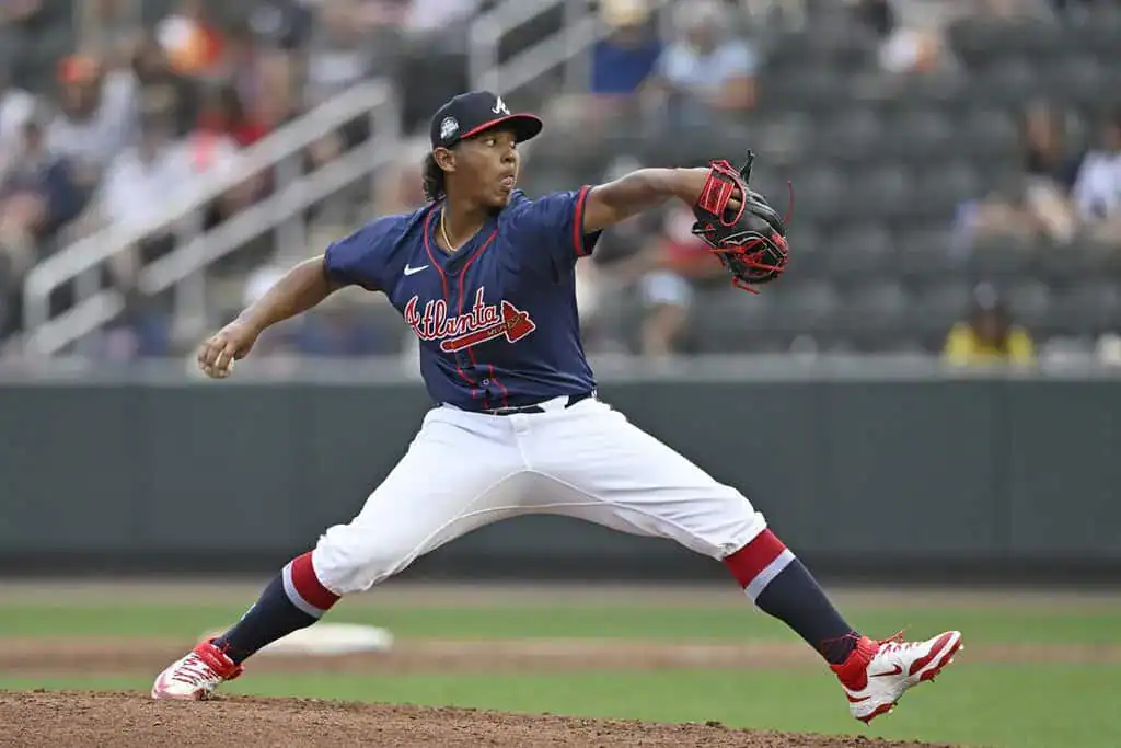 A baseball pitcher in a blue team jersey and white pants throws a pitch on a field, with spectators visible in the background.