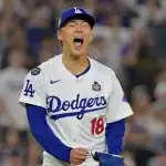 A Los Angeles Dodgers player in a white jersey with blue accents stands on the field, focused during a game night with a cheering crowd behind.