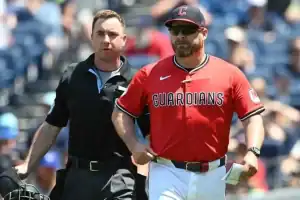 An umpire in a black uniform walks alongside the Cleveland Guardians manager, who is wearing a red jersey, sunglasses, and a blue cap, on a baseball field during a game.