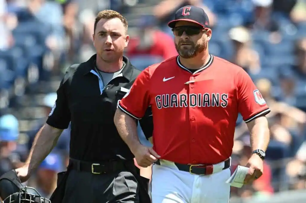 An umpire in a black uniform walks alongside the Cleveland Guardians manager, who is wearing a red jersey, sunglasses, and a blue cap, on a baseball field during a game.