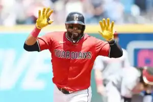 A baseball player in a red Guardians jersey celebrates, raising his hands in excitement during a game on a sunny day.