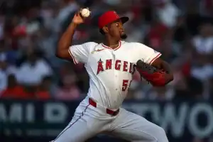 A Los Angeles Angels pitcher in a white jersey winds up to throw a pitch during a game, with a crowd visible in the background.
