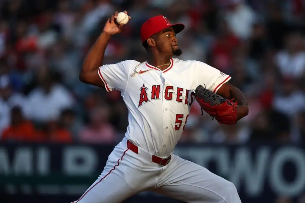 A Los Angeles Angels pitcher in a white jersey winds up to throw a pitch during a game, with a crowd visible in the background.
