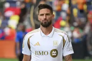 A soccer player in a white jersey with gold accents stands on the field, surrounded by a colorful crowd in the background.